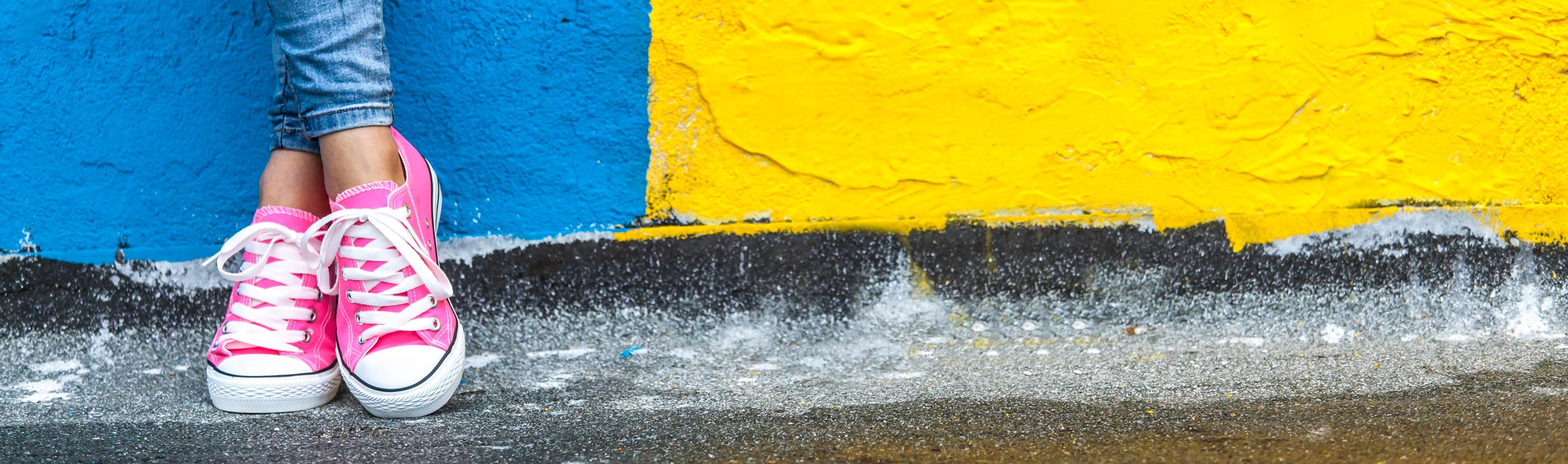 The legs of a girl in blue jeans and pink trainers in front of a blue and yellow wall