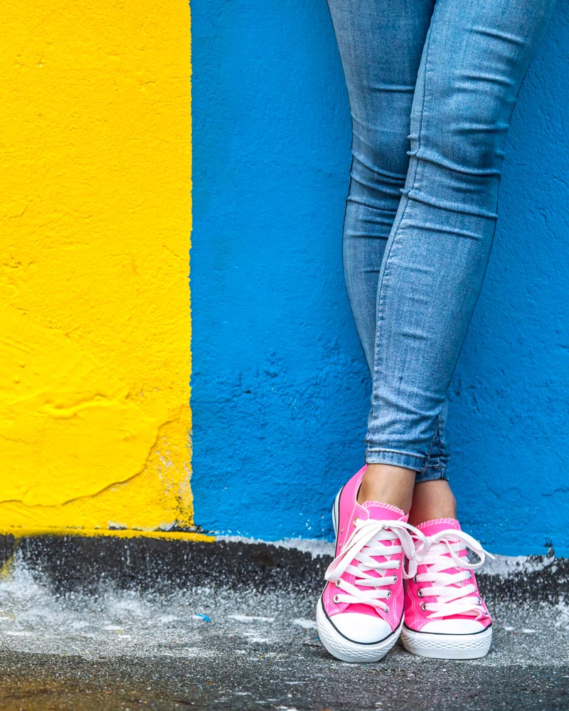 The legs of a girl in blue jeans and pink trainers in front of a blue and yellow wall