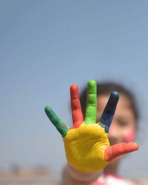 A painted hand held in front of a blurred figure, with the fingers painted different colours and spread out, illustrating the number five