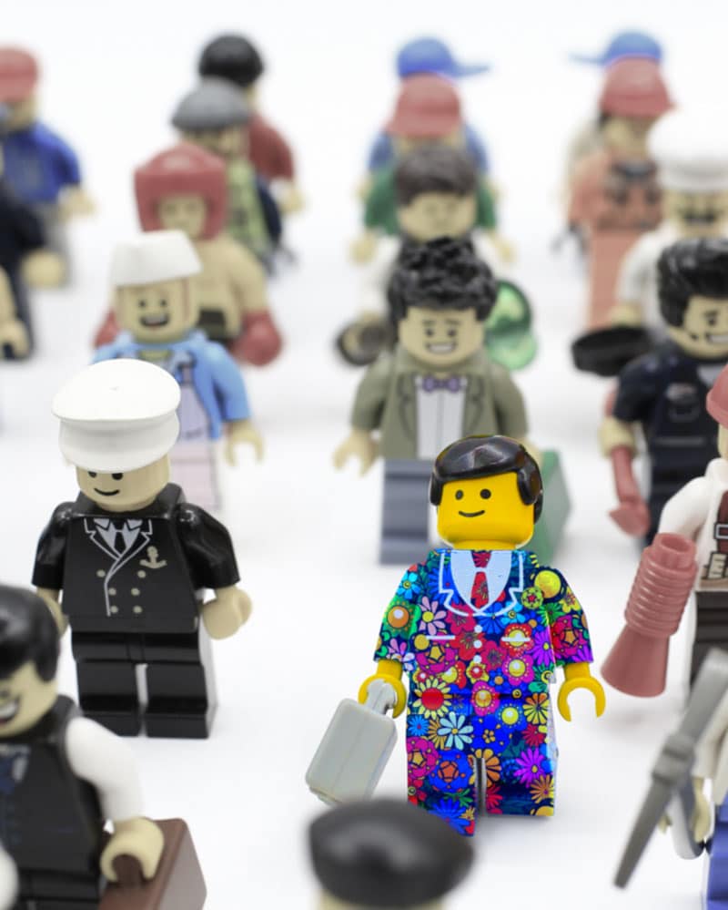 A brightly coloured lego man in a floral suit, standing amongst a crowd of drab lego figures